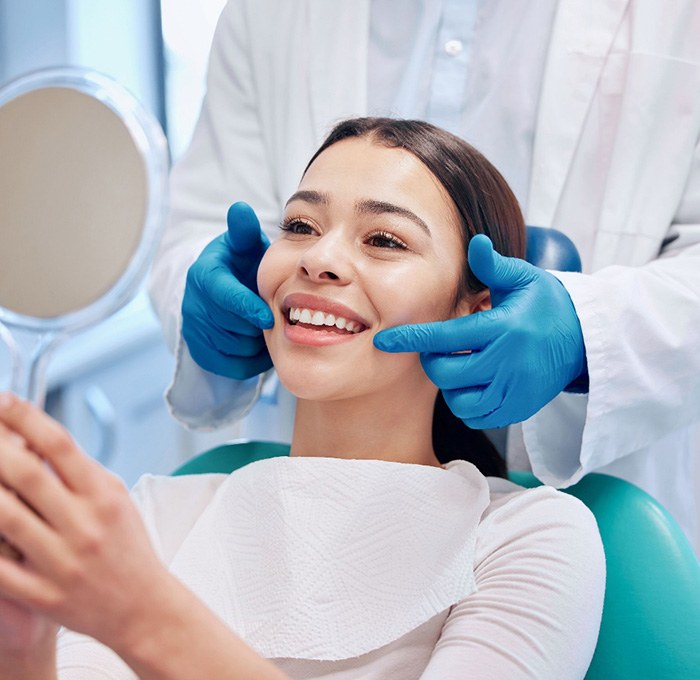 Woman smiling into handheld mirror with dentist’s gloved hands pointing to her smile