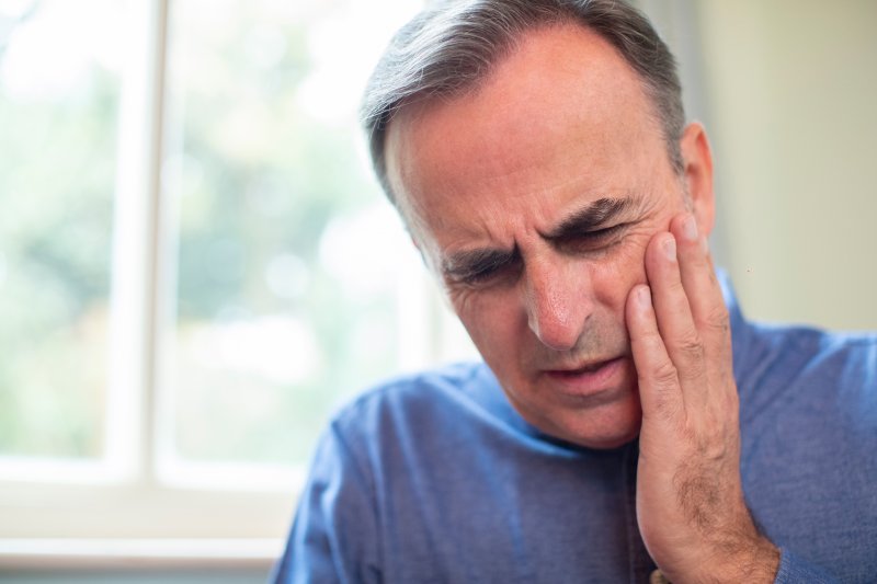 an older man wearing a blue shirt and holding his cheek in pain because of a dental emergency